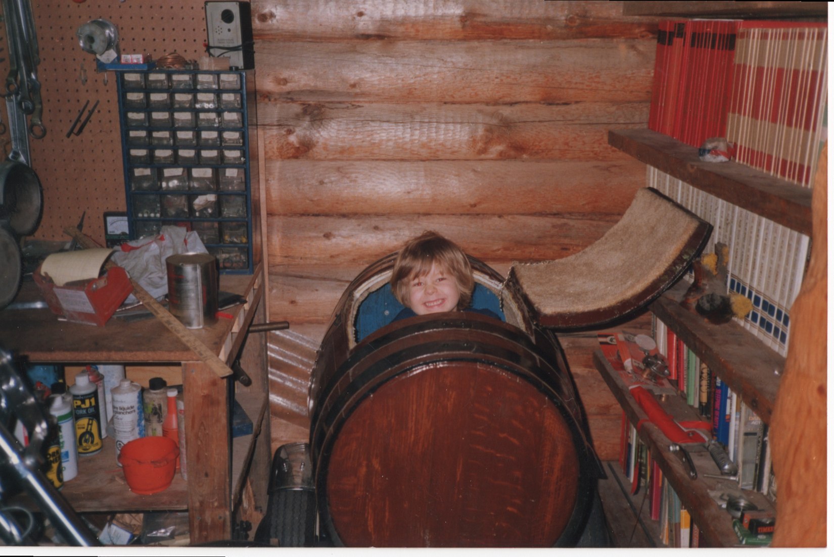 Young child smiling from a wooden barrel in a workshop
